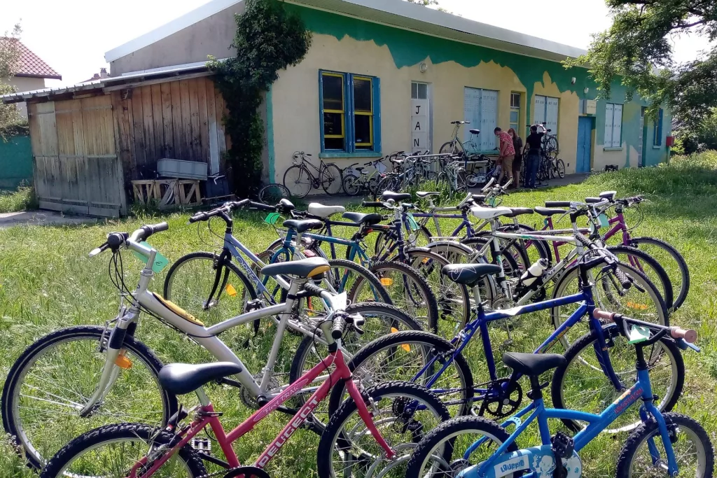 Photo de l'atelier Janus venissieux. Bâtiment vert et jaune aux volets bleu et fenêtres jaunes. Janus est noté tout en longueur sur la porte. Des dizaines de vélos sont exposé dehors sur l'herbe en premier plan. Plus loin devant le bâtiment, deux personne discutent pendant qu'une troisième répare un vélo sur un pied d'atelier.